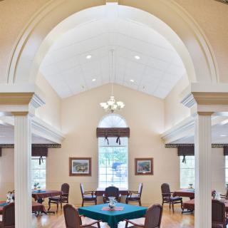 A spacious dining area with an arched ceiling, featuring tables and chairs arranged neatly under a chandelier, surrounded by large windows.