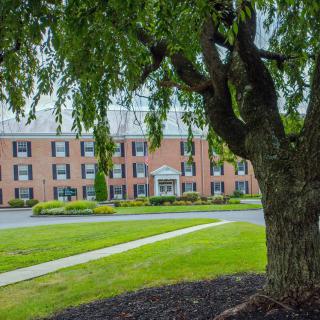 A large brick building with multiple windows, surrounded by well-maintained grass and a prominent tree in the foreground.
