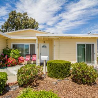 A cozy single-story house with a well-maintained front yard and patio seating, set against a backdrop of blue sky and clouds.