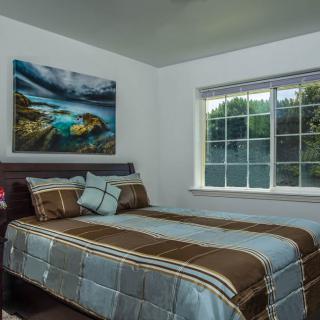 A cozy bedroom with a neatly made bed, a large window, and landscape artwork on the wall.