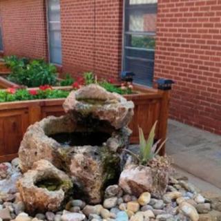 A small rock fountain with surrounding plants in a garden setting against a brick wall.