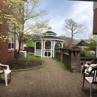 A serene garden with a gazebo and surrounding greenery, featuring a brick pathway and chairs.
