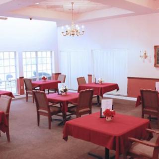 A cozy dining room with several tables covered in red tablecloths, featuring a chandelier and large windows.