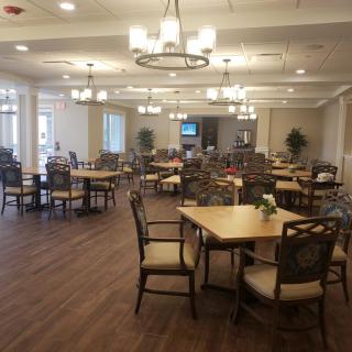 A spacious and elegantly designed dining room with multiple tables and chairs arranged neatly, featuring soft lighting and a wooden floor.