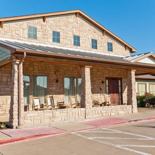 A brick building with a porch and rocking chairs, adjacent to a parking lot on a sunny day.