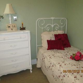 A cozy bedroom with a single white metal bed, adorned with floral bedding and red decorative pillows, alongside a vintage white dresser and lamp.