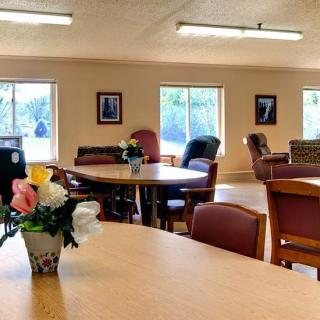 A cozy communal room with tables, chairs, and decorative flowers by large windows.