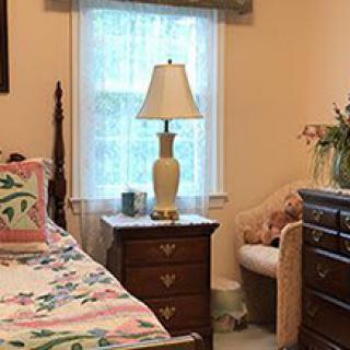 A cozy bedroom with floral bedding, a wooden dresser, and a lamp by the window.