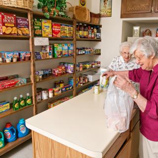 Two elderly women shopping and packing groceries at a small store, surrounded by shelves stocked with various food items.