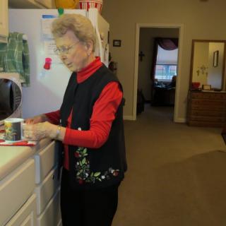 An elderly woman stands in a kitchen with a cup.