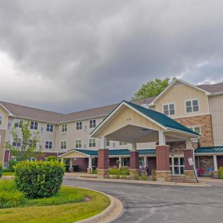 A large residential building with a covered entrance and surrounding greenery under a cloudy sky.