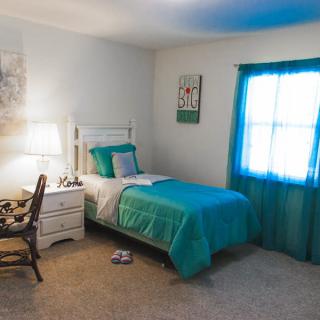 A cozy bedroom with a single bed dressed in turquoise bedding, featuring a nightstand, a lamp, and a window with matching curtains.