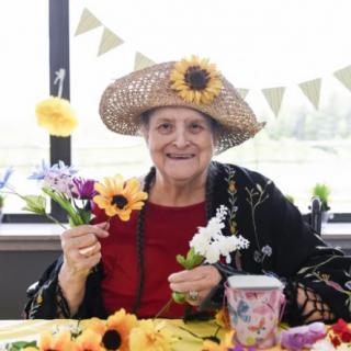 An elderly woman wearing a straw hat adorned with flowers, holding colorful flowers and smiling indoors.