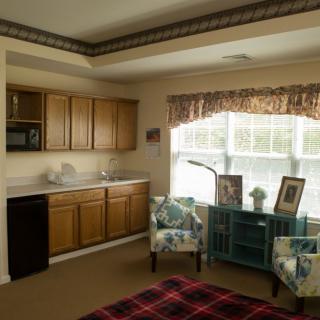 A cozy room with a kitchenette featuring wooden cabinets, two upholstered chairs, and a large window allowing natural light in.
