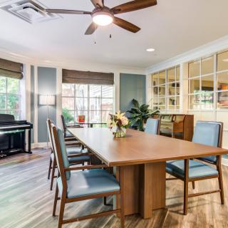 A cozy dining room featuring a large wooden table with chairs, a piano in the corner, and natural light streaming in through the windows.