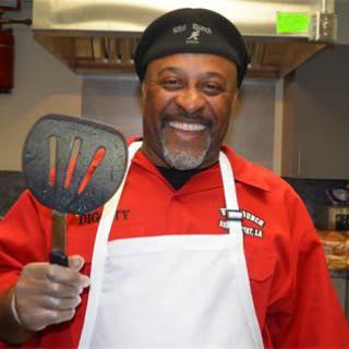A cheerful chef in a red shirt and apron holding a spatula in the kitchen.