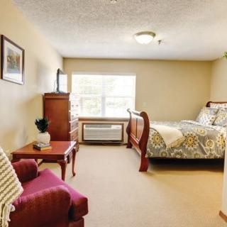 A cozy bedroom with a bed, armchair, wooden dresser, and a large window.