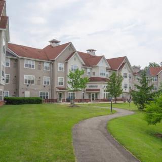 A residential building with a red roof, surrounded by a green lawn and trees, with a pathway leading through the area.