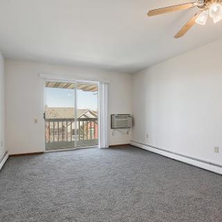 A spacious, empty living room featuring carpet flooring and a ceiling fan, with natural light streaming in from the balcony doors.
