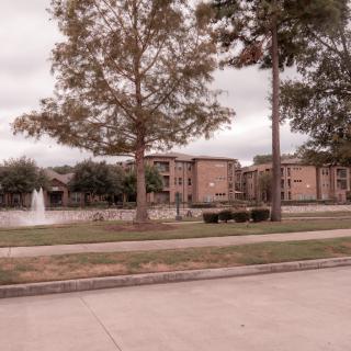 A scenic residential area with apartments by a lake, featuring a fountain and trees under an overcast sky.