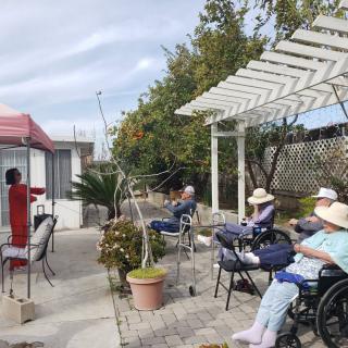 A group of seniors sitting outdoors in a garden setting, some in wheelchairs and wearing hats, listening to a speaker under a pergola.