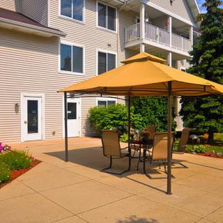 A sunny patio area with outdoor furniture and a large umbrella, adjacent to a building and surrounded by garden landscaping.