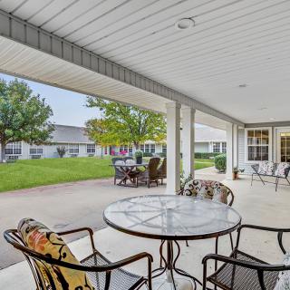 A covered patio with a table and chairs overlooking a lawn and adjacent buildings.