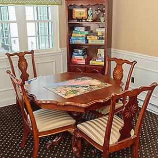 A cozy dining room with a wooden table and chairs, featuring a puzzle in progress and a bookshelf filled with board games and books.