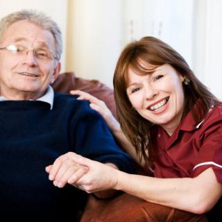 An elderly man and his caregiver smiling while holding hands indoors.