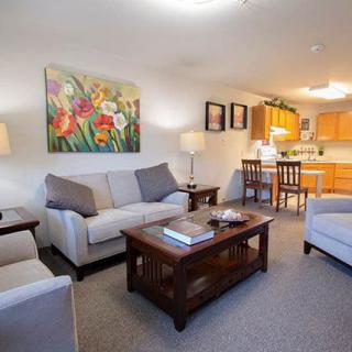 A cozy living room with a sofa set and coffee table, featuring colorful artwork on the wall and an open view of the kitchen area.