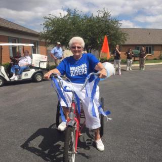 An elderly woman wearing a blue t-shirt rides a bicycle adorned with ribbons during an outdoor celebration.