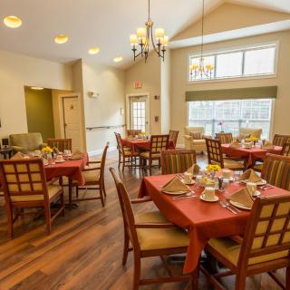 A cozy dining area with neatly arranged tables and chairs, featuring warm lighting and large windows.
