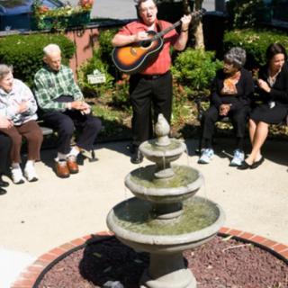 A group of elderly people sitting near a fountain while a man plays the guitar outdoors.