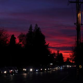 A vibrant sunset casting a colorful glow over the landscape, silhouetted by trees and lined with traffic.