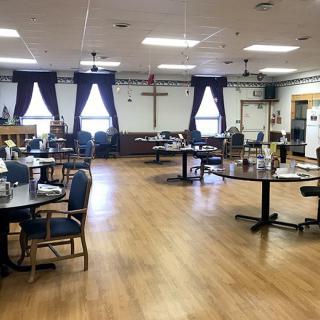 A community dining hall with tables and chairs, featuring a cross on the back wall.