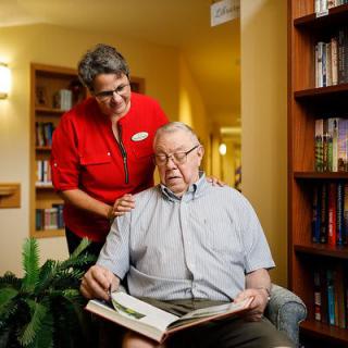An elderly man reading a book with the assistance of a caregiver in a library setting.