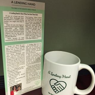 A ceramic mug with 'A Lending Hand' printed on it sits beside a newsletter.