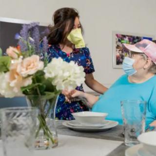 A healthcare worker provides support to an elderly person at a table set for a meal, both wearing masks.