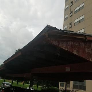 A weathered carport structure with a building in the background under a cloudy sky.