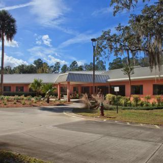 A single-story brick building surrounded by landscaped greenery and trees, with a parking area in front under a blue sky with scattered clouds.