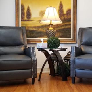 A cozy seating area with two leather chairs, a wooden side table, and a decorative lamp in front of a landscape painting.