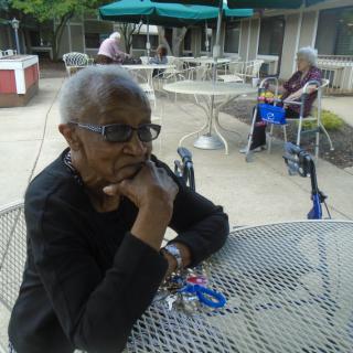 An elderly woman sitting at an outdoor table, looking thoughtful on a sunny day.