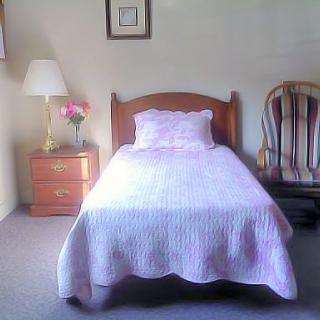 A cozy and simple bedroom featuring a single bed with a pink coverlet, a bedside lamp, and a rocking chair in the corner.