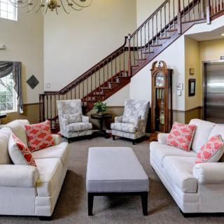 A cozy living room with white sofas and armchairs, decorated with pink cushions, centered around a coffee table, and featuring a grand staircase in the background.