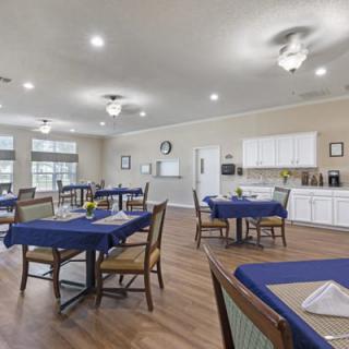 A spacious dining area with wooden floors and tables covered in blue tablecloths, adjacent to a kitchen.