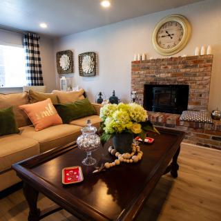 A cozy living room with a comfortable sofa, decorative cushions, a coffee table adorned with flowers, and a brick fireplace featuring a large clock above it.