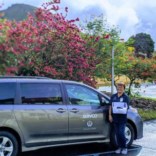A person standing beside a minivan in a scenic outdoor setting with mountains and flowering trees in the background.