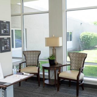 A bright waiting area with two chairs, a table, and a lamp beside large windows.