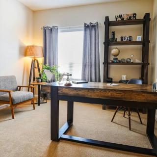 A cozy office space with a wooden desk, chairs, and shelves, illuminated by natural light and a floor lamp.