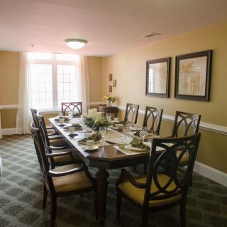 A well-decorated dining room with a long table and elegant chairs, framed by artwork and natural light from the window.